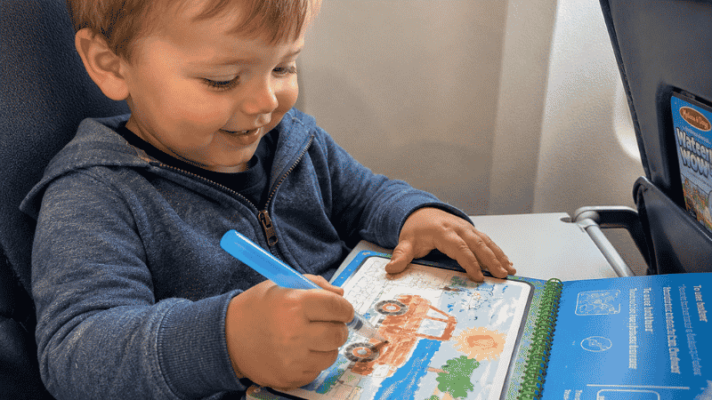 A 3-year-old child using a water brush pen to paint colorful images on a mess-free Water Wow activity book at an airplane tray table